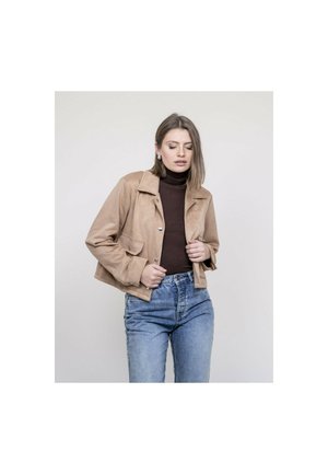 Jeune femme portant une veste beige par-dessus un col roulé marron foncé et un jean bleu, debout devant un fond clair uni.