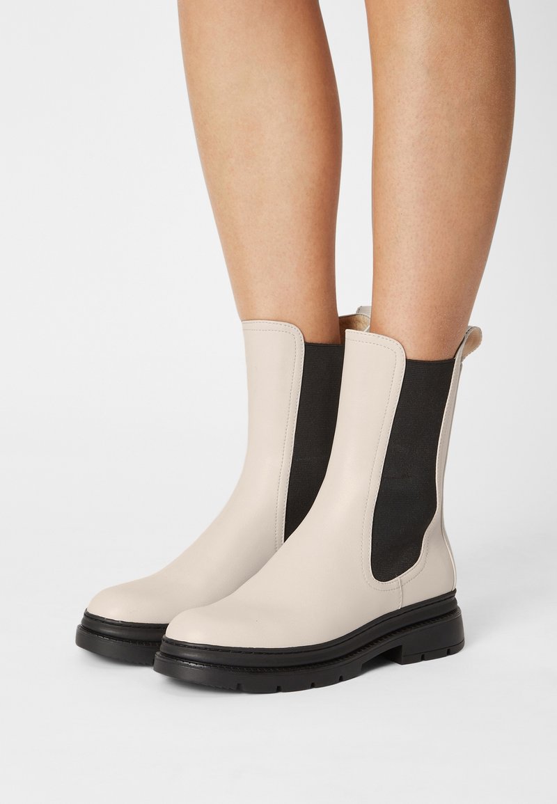 Bottines en cuir beige avec des panneaux élastiques noirs sur les côtés et des semelles épaisses en caoutchouc noir, portées sur des jambes nues.