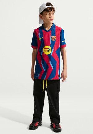 Chłopiec w białej czapce założonej do tyłu, niebiesko-czerwonej koszulce piłkarskiej FC Barcelona z logo Spotify, czarnych spodniach z żółtymi sznurkami oraz czarno-czerwonych trampkach.