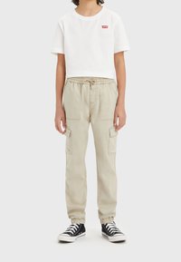 Dziecko ubraniu w białą przylegającą koszulkę Levi's, beżowe spodnie cargo z kieszeniami i wiązaniem w pasie oraz czarne wysokie trampki, stoi na szarym tle.