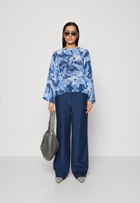 Blusa astratta blu con maniche a campana, abbinata a pantaloni larghi in denim scuro e una borsa a tracolla grigia. Scarpe grigie a punta slip-on.