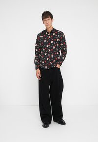 Chemise noire à boutons ornée de motifs floraux colorés et de personnages, associée à un pantalon large noir et des chaussures noires. Coupe et design décontractés.