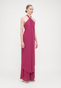 Une robe longue à dos nu en chiffon fuchsia, dotée de drapés doux et d'une coupe décontractée, associée à des sandales à talons blocs blanches.