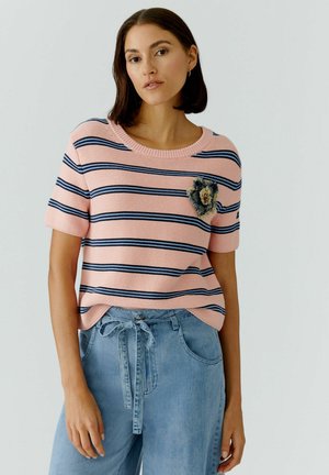 Vrouw draagt een kort mouwtjes trui met roze en marineblauwe strepen, een bloemenpatch en een lichtblauwe high-waisted spijkerbroek met een gestrikte riem.