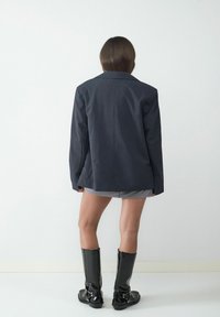 Blazer azul marino oscuro con corte entallado y solapas con muesca, combinado con pantalones cortos gris y botas negras brillantes hasta la rodilla.