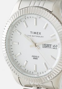 Montre en acier inoxydable argenté avec un cadran blanc, index et aiguilles argentés. Dispose d'un affichage de la date et d'une lunette texturée et striée.