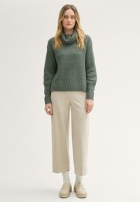 Zielony sweter dziany z dekoltem typu cowl oraz ściągaczem u dołu, w połączeniu z jasnobeżowymi spodniami o szerokim nogawce i kremowymi skarpetami, noszony z jasnymi butami.