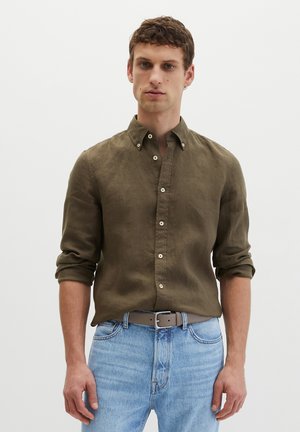 Uomo che indossa una camicia verde oliva con bottoni e maniche arrotolate e jeans azzurri con cintura grigia su sfondo bianco uniforme.