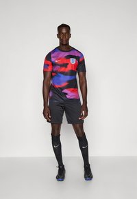 Camiseta de fútbol Nike con base negra y patrones abstractos en rojo y azul, mangas cortas y escudo de Inglaterra. Pantalones cortos y calcetines negros.