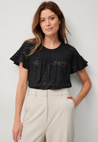 Schwarze Bluse mit Ösen und Rüschenshortsärmeln, die einen taillierten Oberkörper und aufwendige Ausschnittmuster aufweist. Kombiniert mit hellbeigen Hosen.
