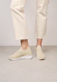 Des baskets en maille beige dotées d'un design respirant, d'un style slip-on et d'une semelle blanche épaisse avec un léger talon. Accents texturés tout au long.