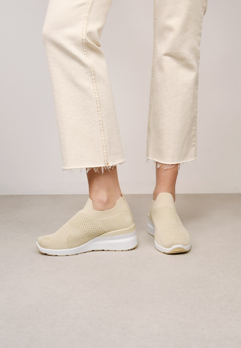 Des baskets en maille beige dotées d'un design respirant, d'un style slip-on et d'une semelle blanche épaisse avec un léger talon. Accents texturés tout au long.