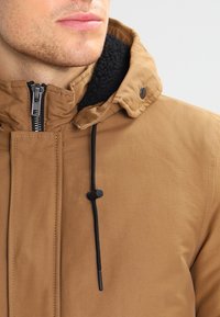 Brown zip-up jacket with a textured black lining, adjustable drawstring hood, and snap button accent on the collar. Smooth fabric finish.