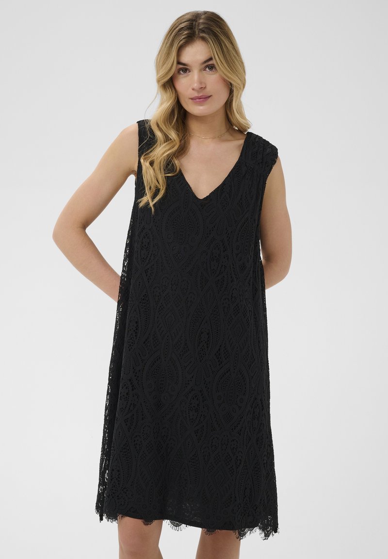 Femme portant une robe noire en dentelle sans manches avec un décolleté en V, debout, une main dans le dos, sur un fond gris uni.