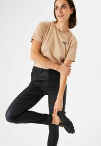 Camiseta de algodón beige con un estampado de tigre negro, combinada con pantalones ajustados negros. El modelo posa con un pie levantado, mostrando calzado negro.