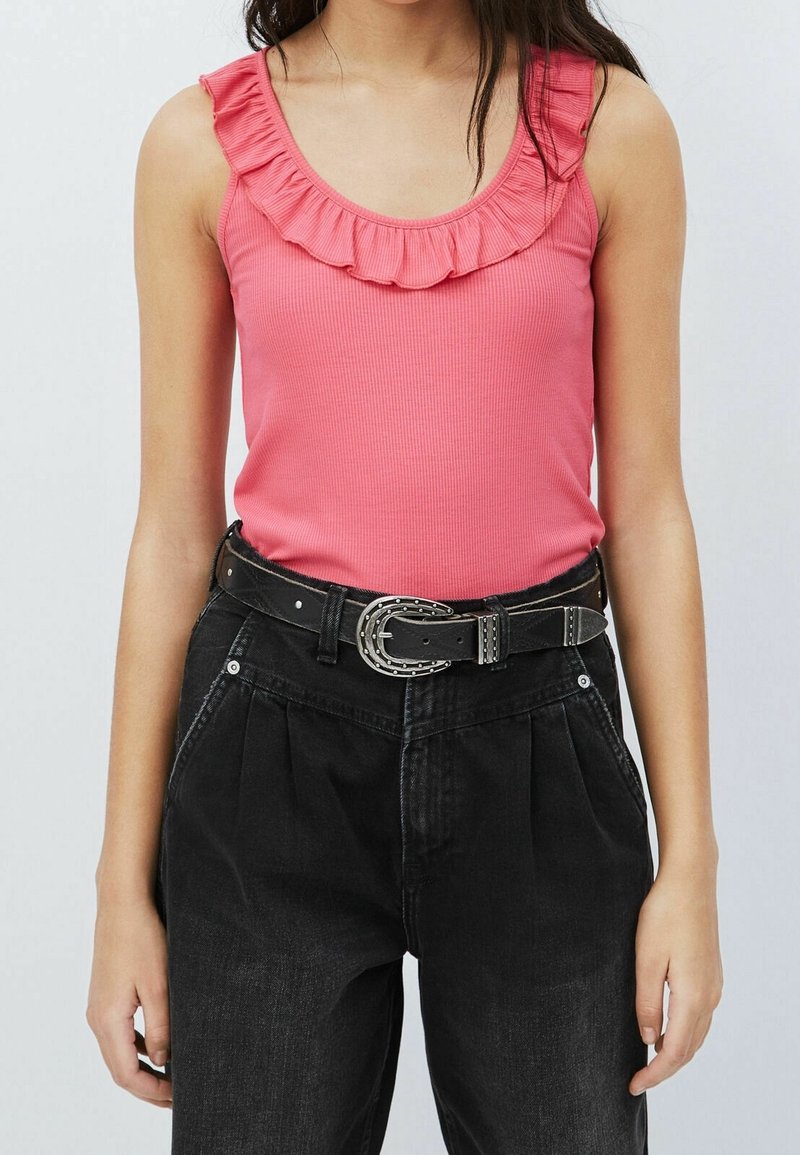 Débardeur rose à côtes avec un col à volants, associé à un pantalon noir taille haute et une ceinture noire avec une boucle en argent et des clous.