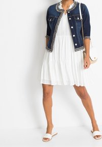 Denimová bunda s odstřiženými okraji, bílá šaty s volány a bílé skluzné sandály. Oblečení obsahuje tašku přes rameno a knoflíkové detaily.