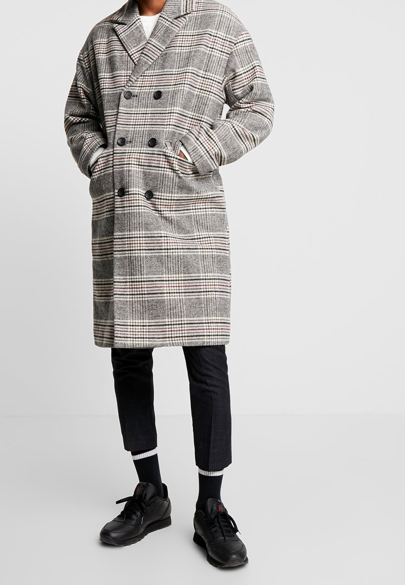 Double-breasted plaid coat in grey with red accents, featuring large buttons and side pockets. Paired with black pants and shoes.