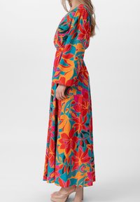 Robe longue colorée avec imprimé floral en rouge, orange et teal ; manches bouffantes et taille cintrée ; portée avec des talons compensés beiges.