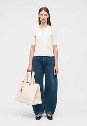 Γυναίκα όρθια φορώντας λευκό μπλουζάκι με κοντά μανίκια και γιακά, μπλε τζιν παντελόνι φαρδύ, μαύρα φλατ παπούτσια, κρατώντας μεγάλη κρεμ τσάντα ώμου με καφέ λεπτομέρειες.