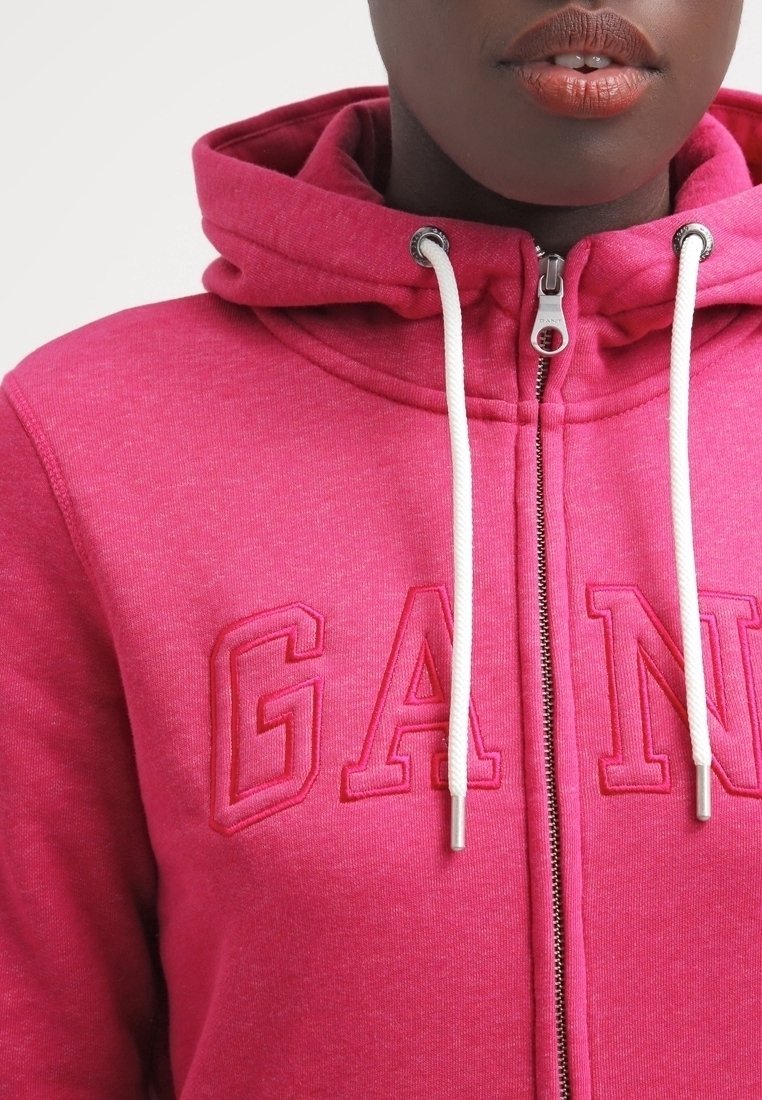 Roze zip-up hoodie met een textuurstof, grote geborduurde "GANG" letters, witte koorden en een trekkoord capuchon.