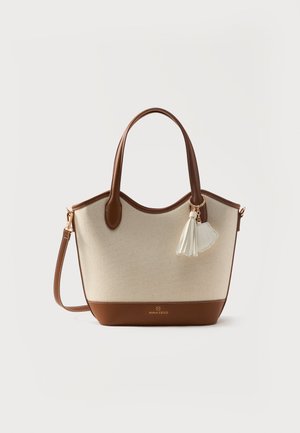 Sac fourre-tout en toile beige avec anses et garnitures en cuir marron, bandoulière amovible, et un pompon blanc ainsi qu'un charm en forme de coquillage.