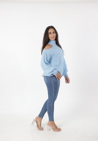 Mujer con cabello largo y oscuro, llevando un suéter azul claro con cortes en los hombros, jeans ajustados azules y sandalias de tacón transparente, de pie de lado.