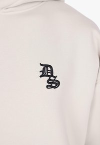 Detailný záber na svetlo béžovú mikinu s čiernymi vyšívanými písmenami v gotickom štýle "DS" na hrudi.