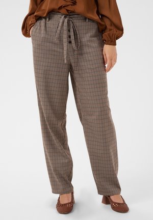 Femme portant un pantalon marron à carreaux avec une ceinture nouée à la taille, un chemisier marron à volants sur le devant et des chaussures marron tressées à talon carré.
