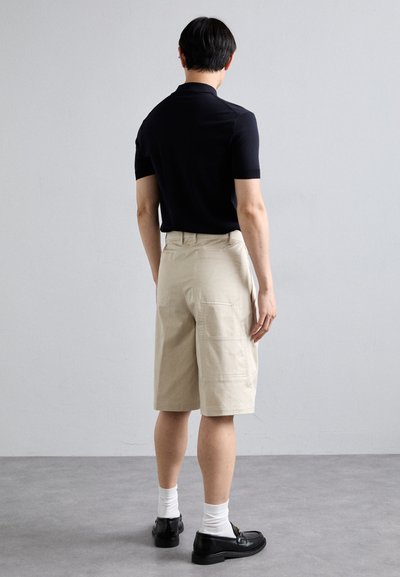 Shorts cargo beige avec poches latérales, associés à un polo noir à manches courtes et des mocassins noirs, portés avec des chaussettes blanches.