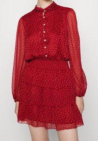 Röd lagrad klänning med knäppning fram, genomskinliga långärmar, hög hals och blommönster med små vita och blå detaljer.