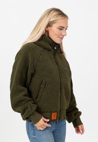 Zielona oliwkowa polarowa kurtka z wysokim kołnierzem, zamkiem błyskawicznym i prążkowanymi mankietami. Posiada boczne kieszenie oraz teksturowany materiał z pomarańczową metką.