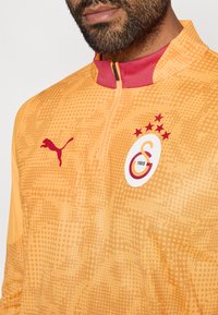 Pomarańczowy sportowy polar z wysokim kołnierzem, z czerwonym zamkiem i akcentami. Teksturowany wzór, logo Pumy oraz emblemat drużyny z gwiazdami.