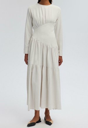 Longue robe crème avec taille froncée et volants sur la jupe, manches longues, portée avec des ballerines pointues noires et beige.