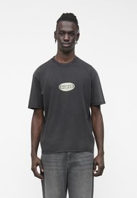 Čierne bavlnené tričko s okrúhlym výstrihom a krátkymi rukávmi. Na prednej strane je potlačené zelené logo "GUESS". Nosené so sivými džínsami.