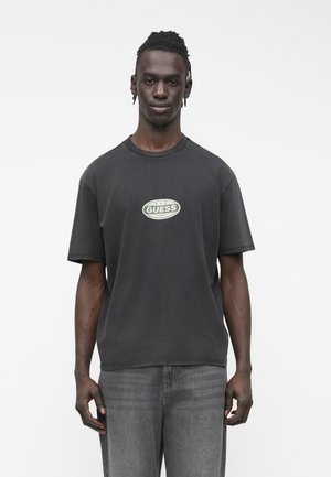 Melna kokvilnas T-krekls ar apaļu izgriezumu un īsām piedurknēm. Priekšpusē zaļā krāsā uzdrukāts "GUESS" logotips. Valkāts kopā ar pelēkām džinsa biksēm.