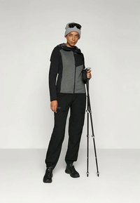 Person in gray beanie, black-gray jacket, black pants, hiking shoes, holding folded trekking poles, standing against plain background.