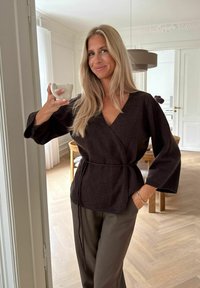 Dunkelbrauner Wickelpullover mit weiten Ärmeln, in der Taille gebunden, kombiniert mit olivgrünen Hosen, während sie eine kleine hellfarbige Tasse hält.