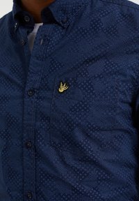 Tmavě modrá košile na knoflíky s texturovaným vzorem, s malým žlutým vyšitým logem na náprsní kapse a tmavými knoflíky.