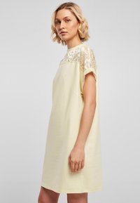 Short-sleeved, knee-length dress in pale yellow cotton with a lace overlay at the shoulders, featuring a round neckline and relaxed fit.
