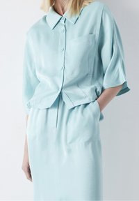 Camisa de botón de manga corta y color azul claro, con cuello y un bolsillo en el pecho, fabricada en un tejido suave, combinada con pantalones de tiro alto a juego.