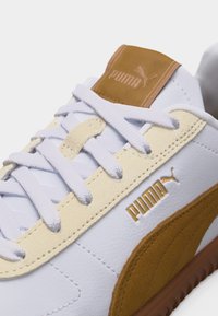 Gros plan d'une basket blanche Puma avec des détails en daim beige, des lacets blancs et un logo Puma doré sur le côté et la languette.