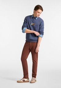 Marineblauer Pullover mit Dackelgrafik, über einem hellblauen Shirt. Braune Slim-Fit-Hosen und beiges Sneakers mit grünen Akzenten.