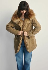 Brązowa parka z kołnierzem z sztucznego futra, z zamkiem z przodu, guzikami i kieszeniami po bokach, w zestawie z niebieskimi dżinsami.