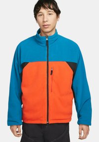 Fleecejacka med hög krage, med en blå övre del, orange nedre del, svarta accenter och en dragkedjeficka på bröstet.