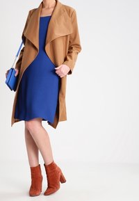 Cappotto marrone a maniche lunghe con colletto drappeggiato sopra un vestito blu. Stivaletti in suede rossi alla caviglia con tacco spesso. Piccola borsa blu con tracolla a catena.