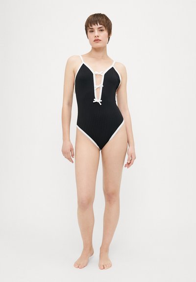 Maillot de bain une pièce noir à côtes avec des bordures blanches, décolleté en V profond et détails à lacets à l'avant. Texture douce, design élégant.