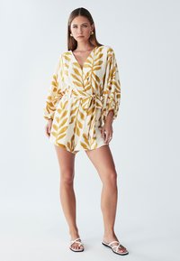 Witte romper met geel bladprint, overslagontwerp, lange mouwen en een ceintuur in de taille. Model draagt witte slippers. Gladde stofafwerking.