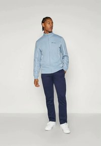 Veste zippée bleu clair avec des garnitures blanches, associée à un pantalon bleu marine et des chaussures blanches. Le tissu semble lisse et présente un design décontracté.