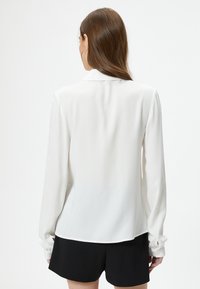 Blusa bianca a maniche lunghe con colletto, texture liscia, vestibilità leggermente ampia e polsini abbottonati, abbinata a pantaloni corti neri.